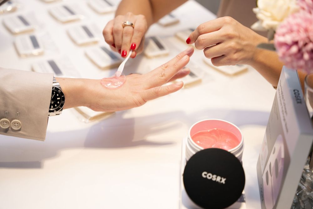 A beauty consultant applies COSRX pink cream product on a customer's hand at a cosmetic display counter.