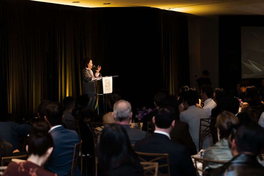 Speaker presenting at a formal conference event with an attentive audience seated in a darkened ballroom.