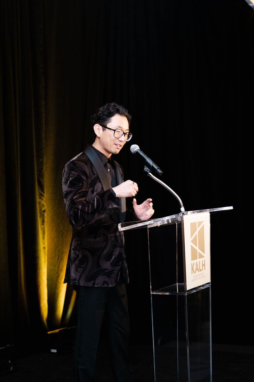 Man in patterned blazer speaking at KALH podium on stage with microphone at formal event.