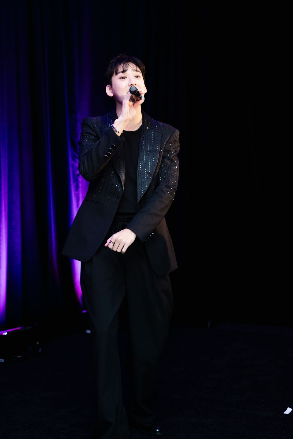 A performer in a black suit sings into a microphone on a dimly lit stage with purple lighting.