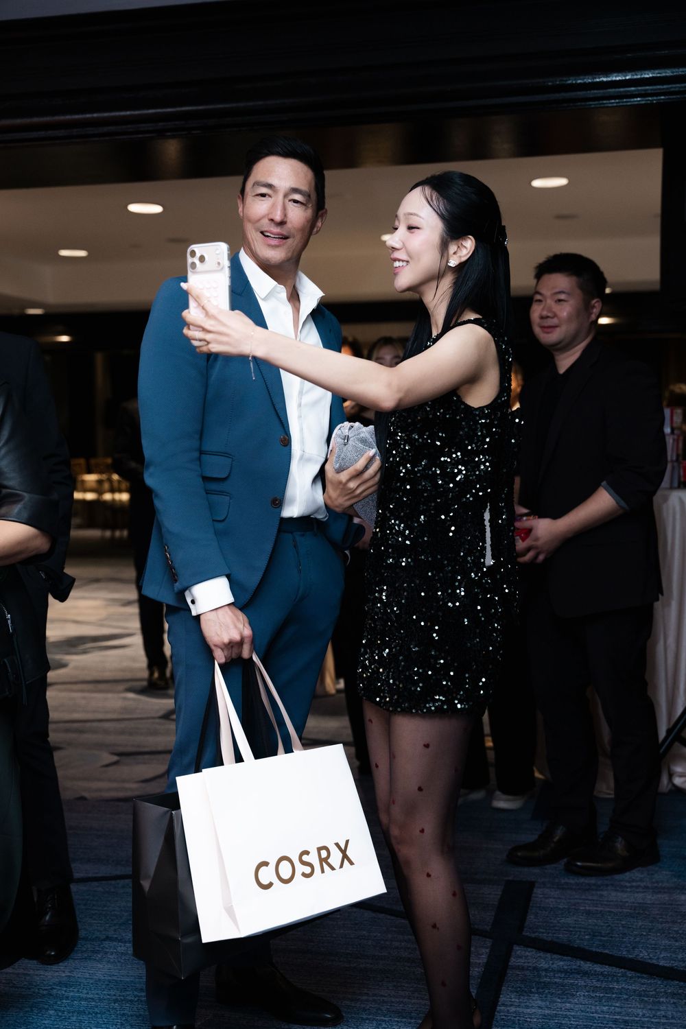 Two elegantly dressed guests take a selfie at a COSRX event, man in blue suit holding branded shopping bag.