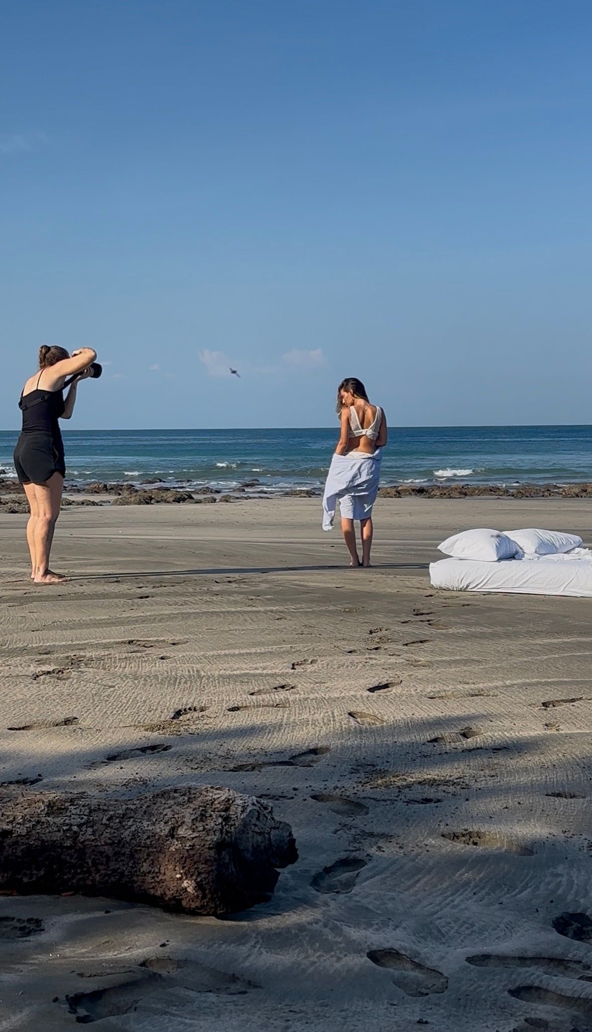 Photographer capturing a model wrapped in white fabric during a beach photoshoot on a sunny day.