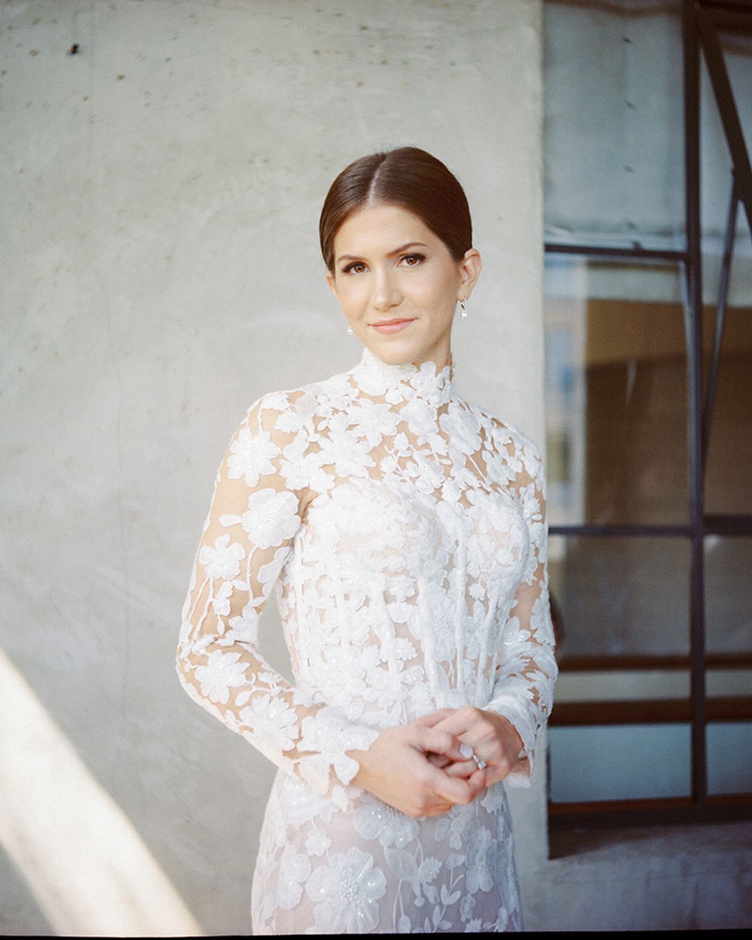 A woman in a delicate white lace dress poses against a light wall in a soft, dreamy bridal portrait series.