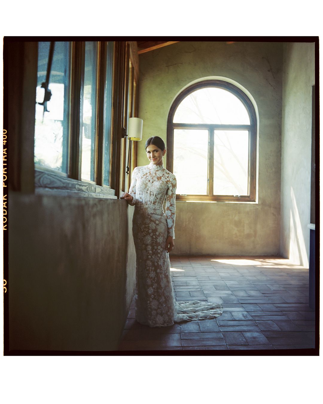 A person in a floral dress stands in a sunlit stone hallway with arched windows and rustic flooring.