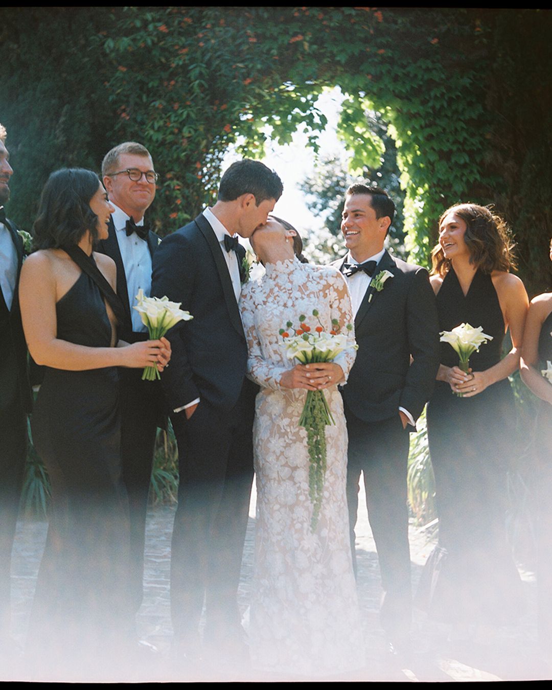 Wedding party lineup outdoors with guests in formal attire holding bouquets against a bright natural backdrop.