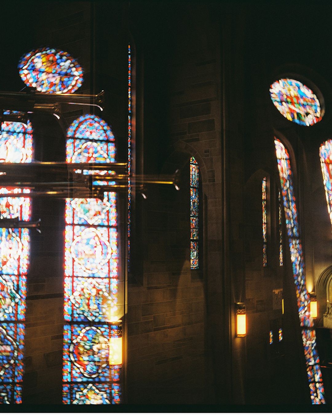 Stained glass windows cast colorful light through dark gothic church interior with stone columns and arches.
