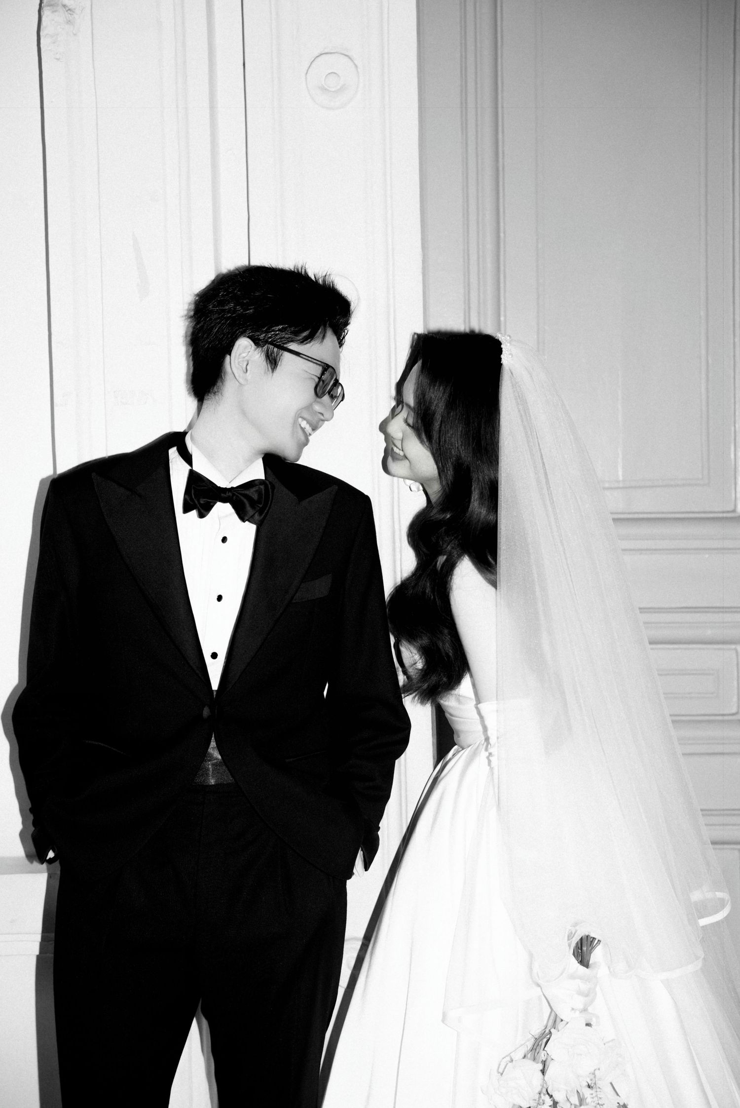 Black and white wedding portrait of smiling groom in tuxedo and bride in veil against white wall.