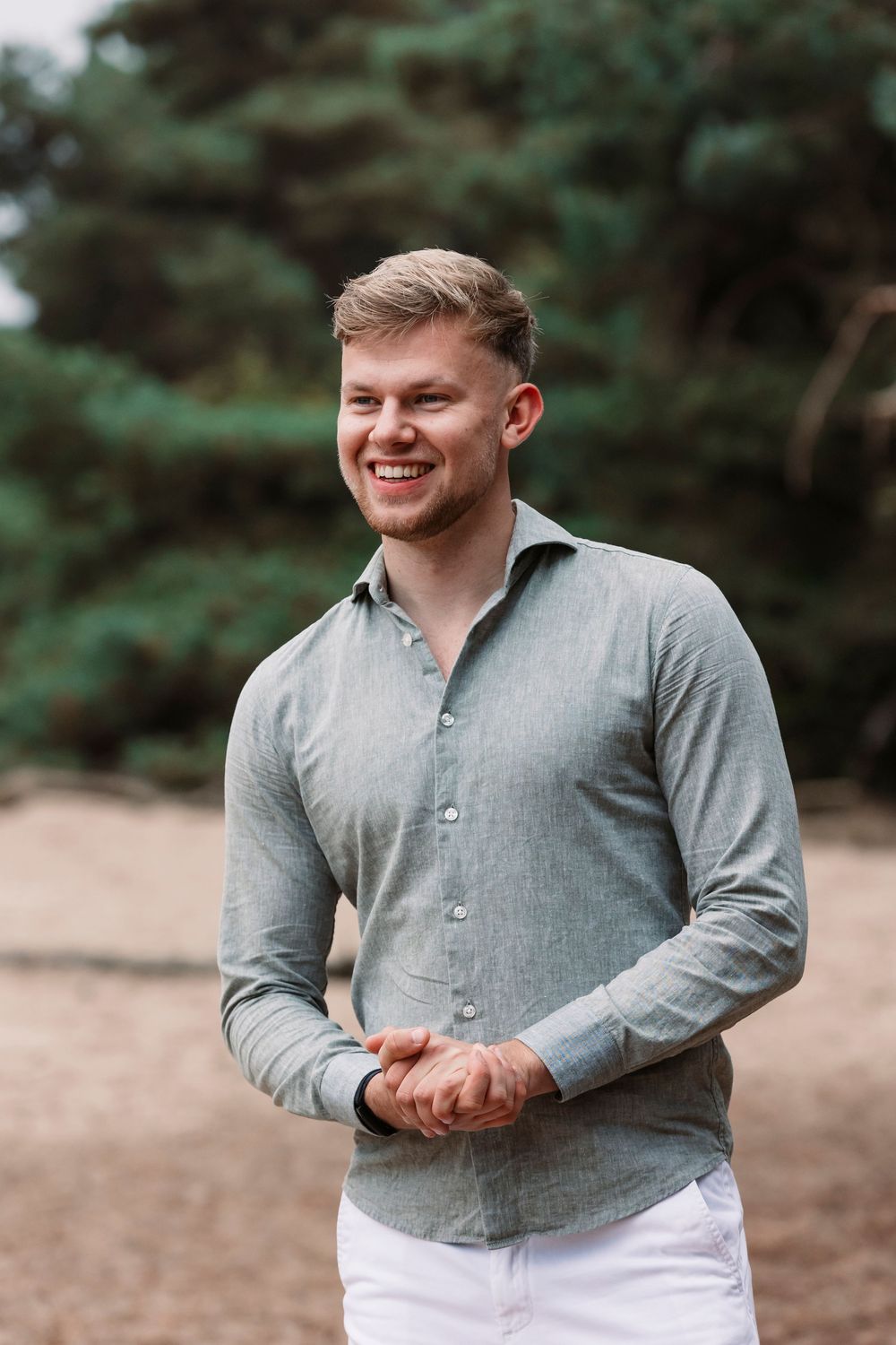 Cheerful outdoor portrait showing person in gray shirt and white pants against pine trees.