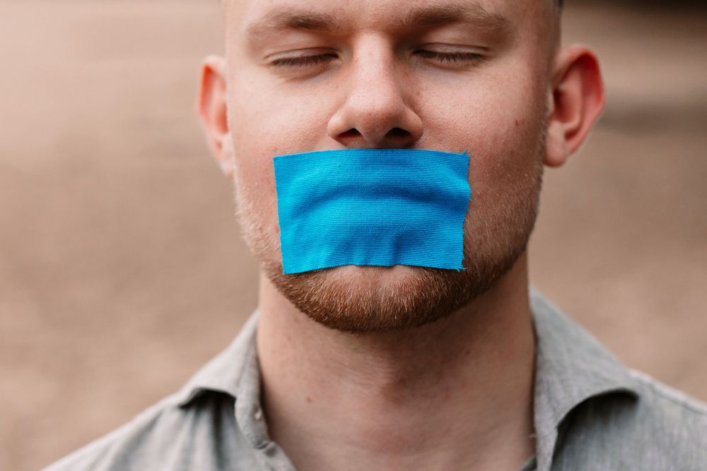 Creative portrait showing person with bright blue tape over mouth against natural background.
