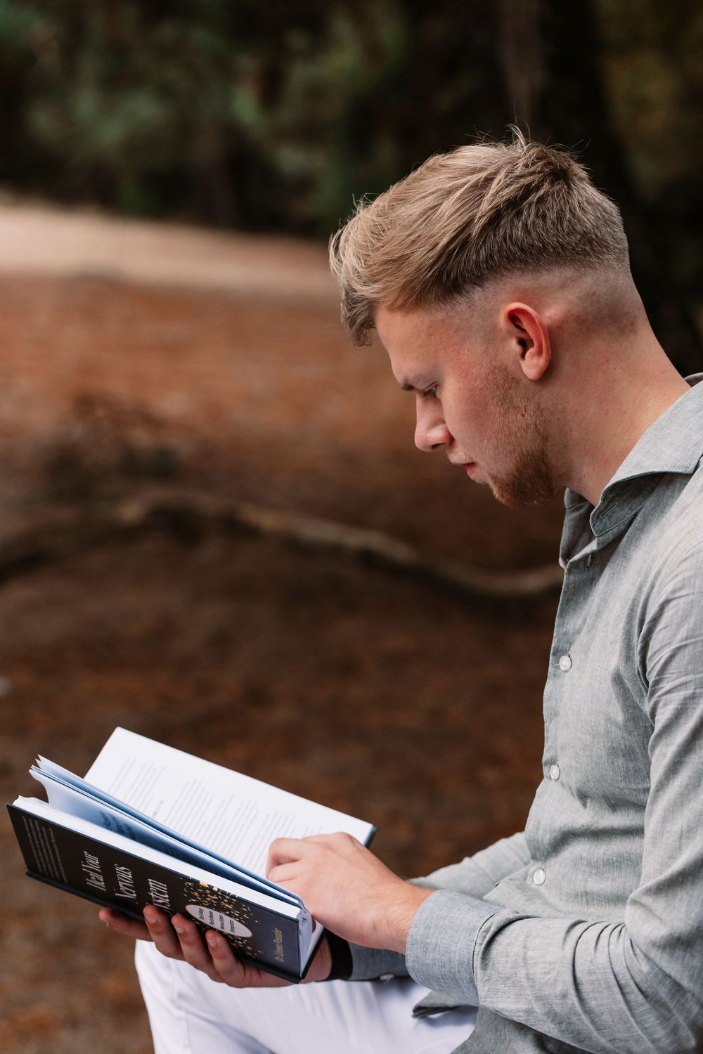 Person in gray shirt reading book while sitting outdoors in natural setting.