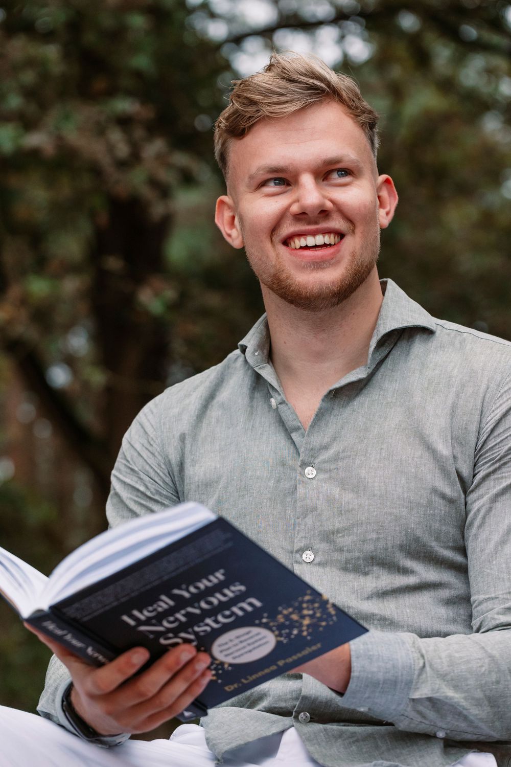 Person smiling while reading book outdoors, wearing gray casual shirt against forest background.