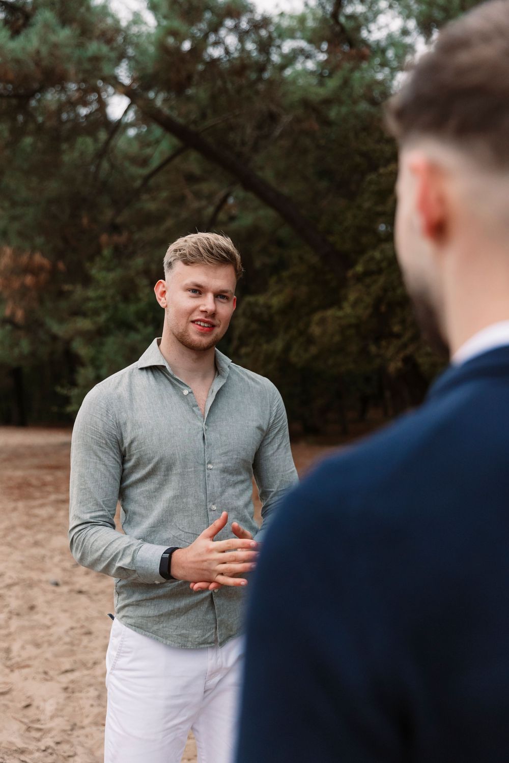 Person in grey shirt gestures animatedly during outdoor conversation in wooded setting.