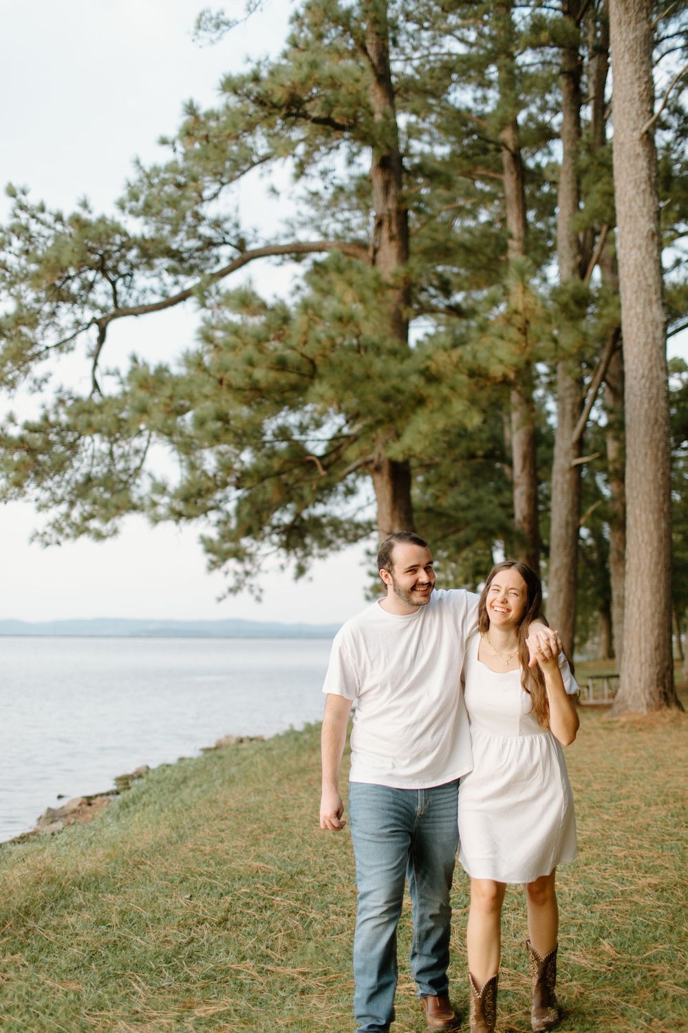 Romantic couple walking along a scenic lakeside path with tall pine trees.