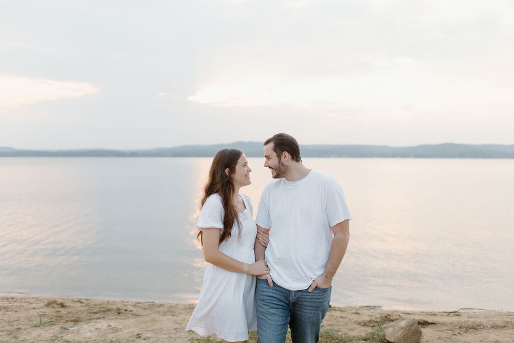 Couple in white clothing embracing by the waterfront during sunset.