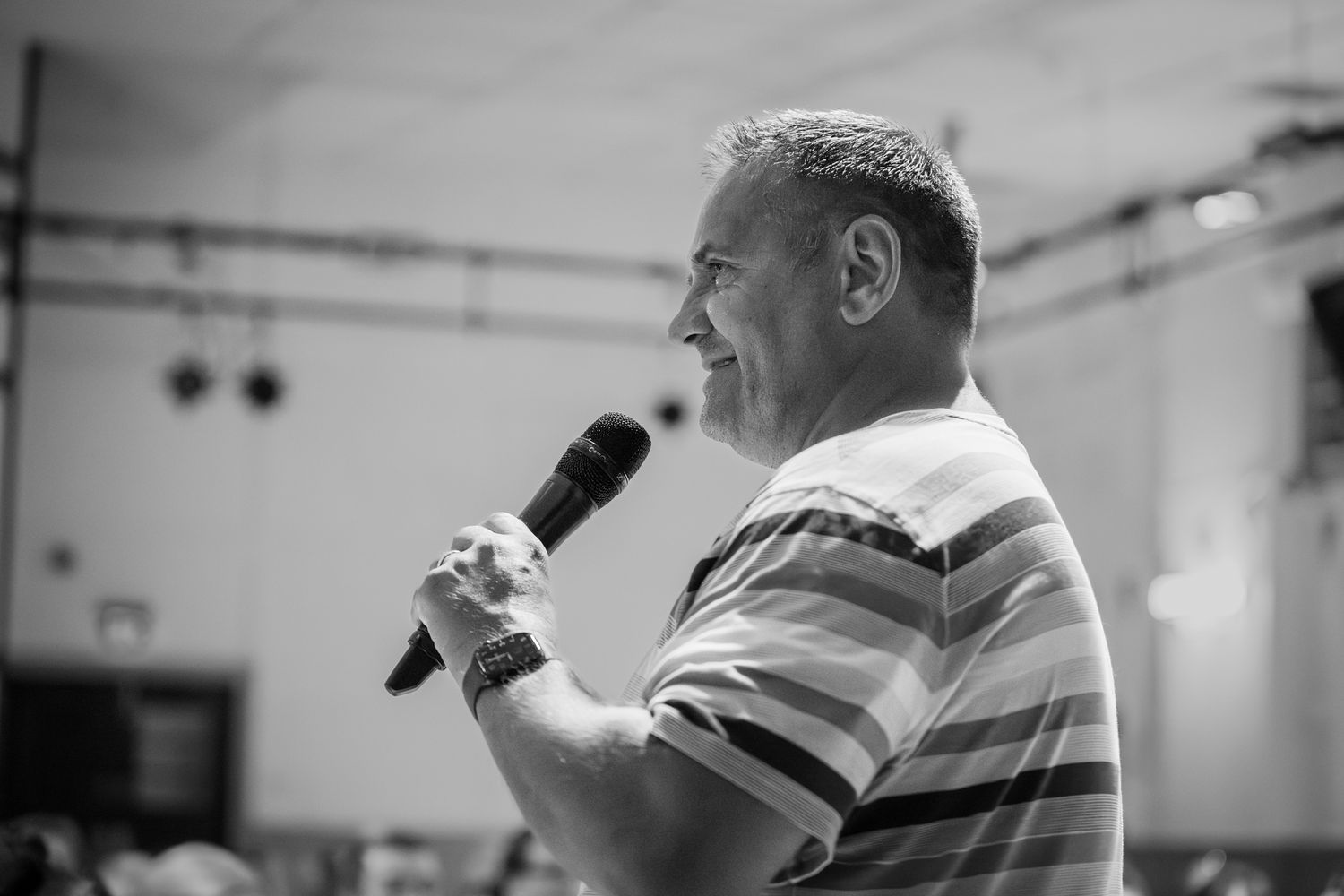 Black and white photo of a smiling man speaking into a microphone at an indoor event.