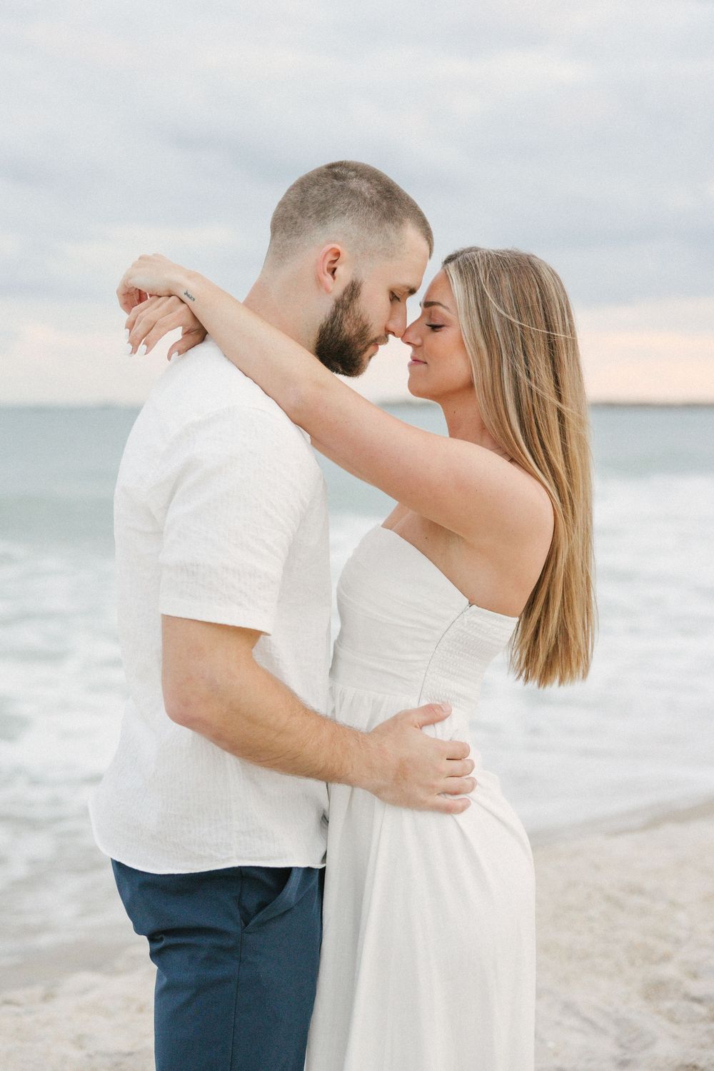 A romantic couple shares an intimate moment dancing together on a beach at sunset in matching white outfits.