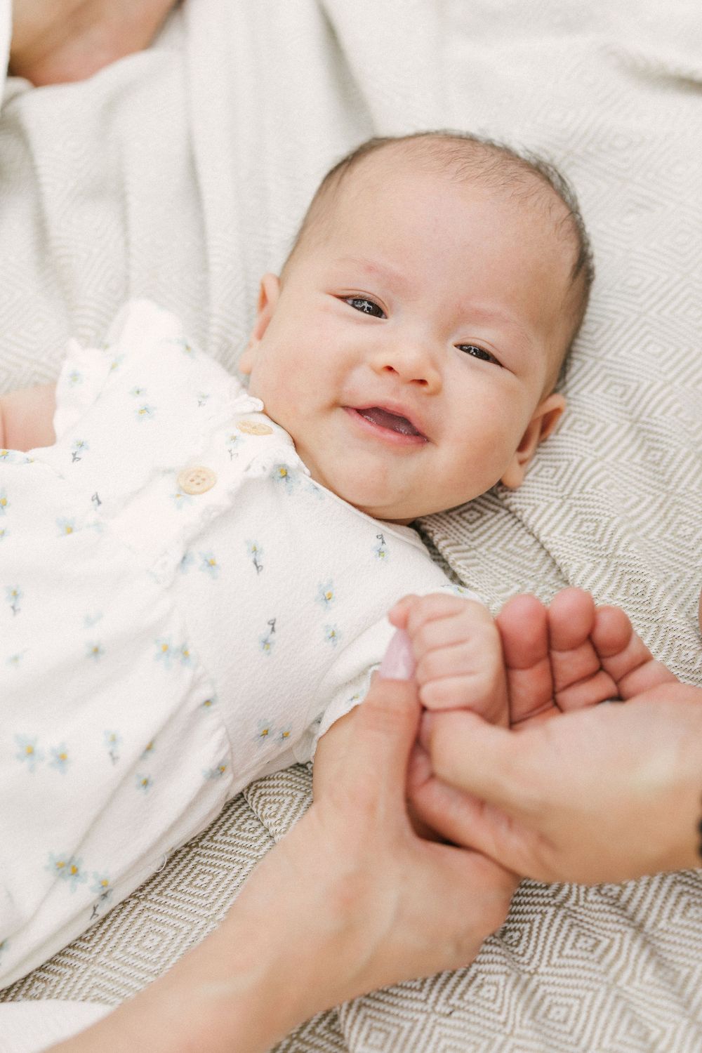 A smiling baby in white clothing lies on a gray and white patterned blanket while holding an adult's hand.