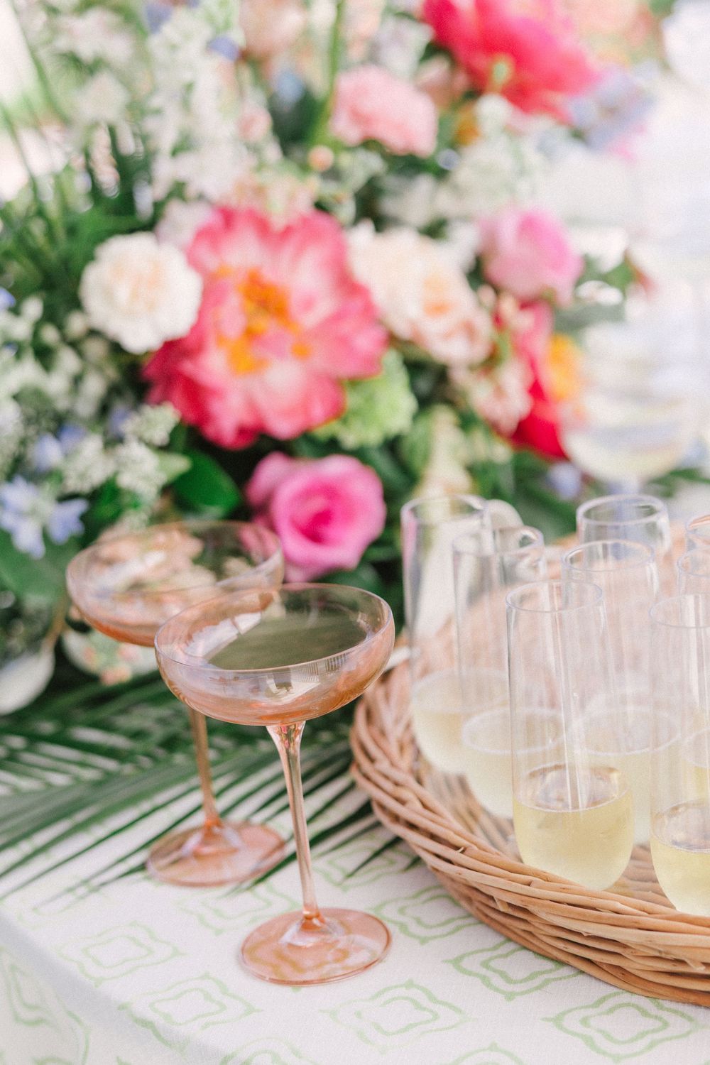 Pink champagne coupe glasses and clear cocktail glasses arranged on wicker chargers with coral flowers in background.