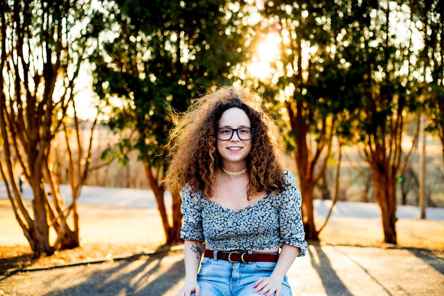 A curly-haired photographer in glasses poses outdoors during golden hour sunset.
