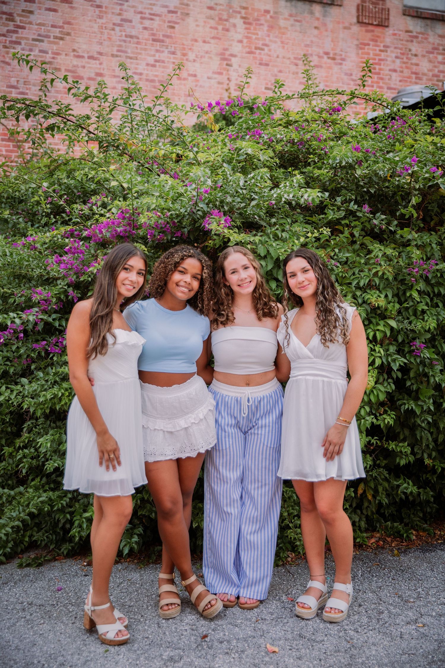 A group wearing light summer outfits poses together against a flowering bush backdrop on a gravel path.