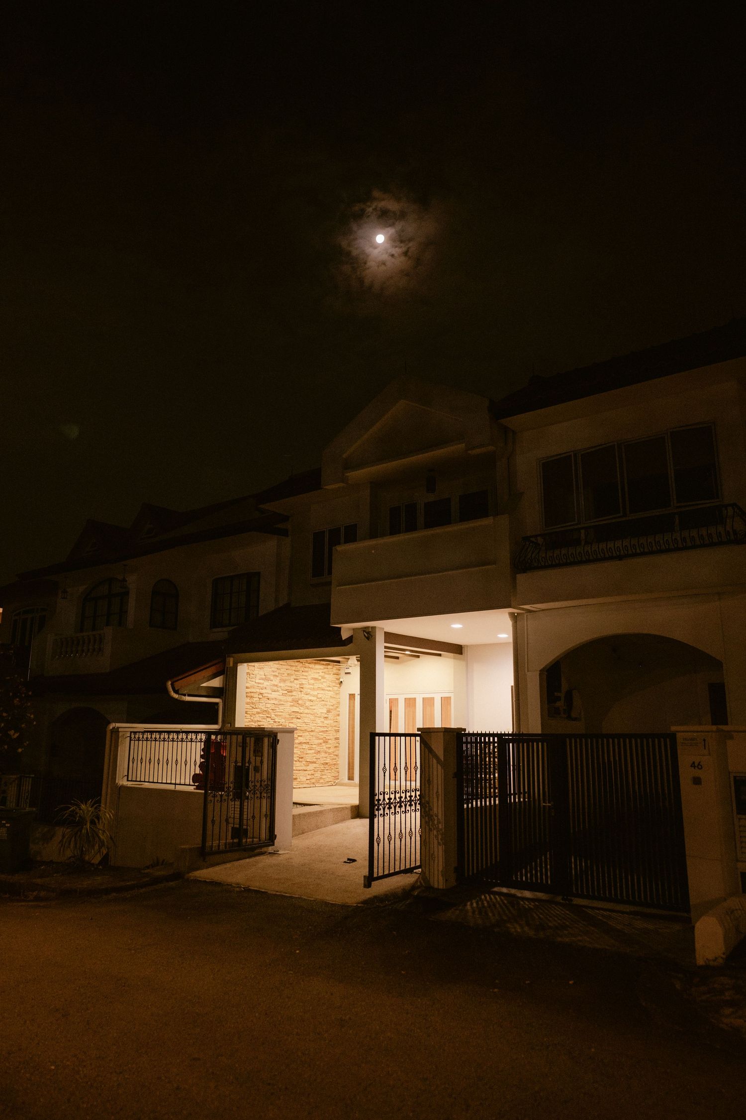 Moonlit night sky over a two-story residential house with illuminated entrance and iron gate.