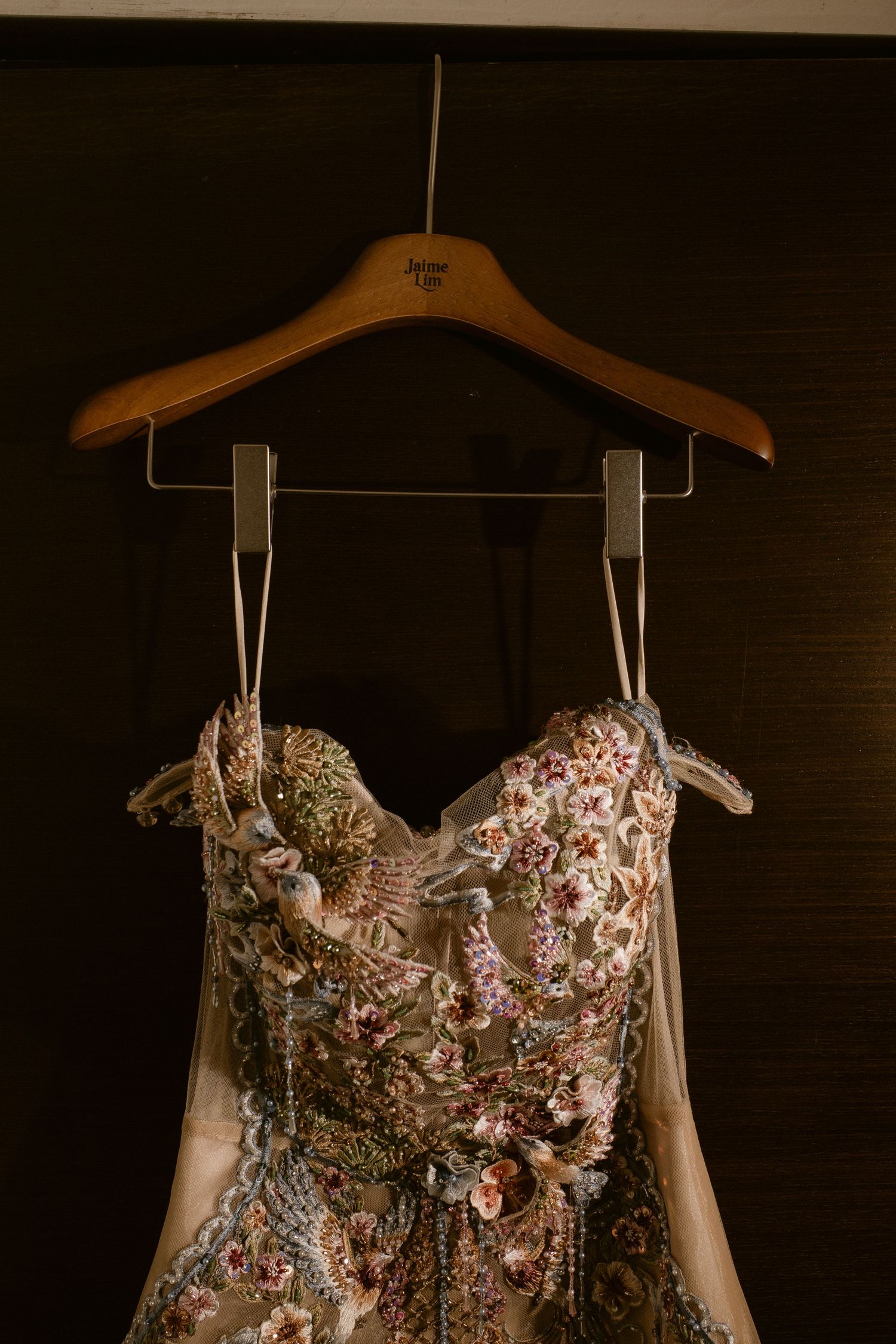 Intricate floral embroidered wedding gown hanging on a wooden hanger against a dark background.