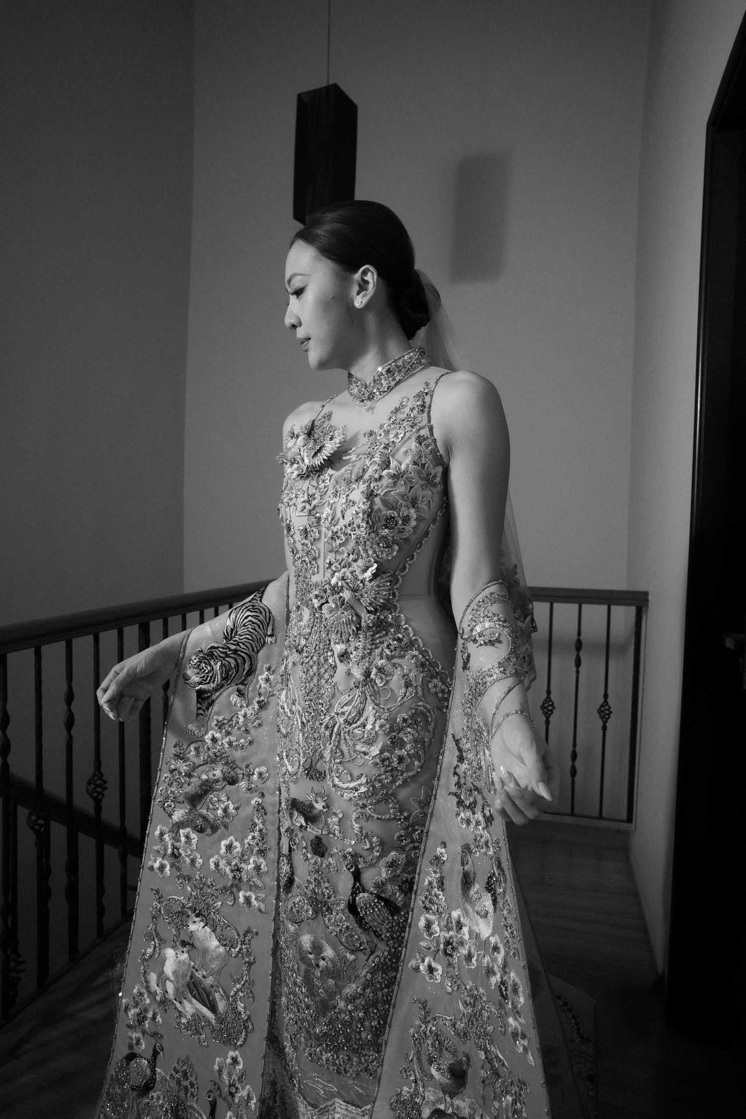 Black and white portrait of a bride in an ornate embroidered gown standing near a staircase.
