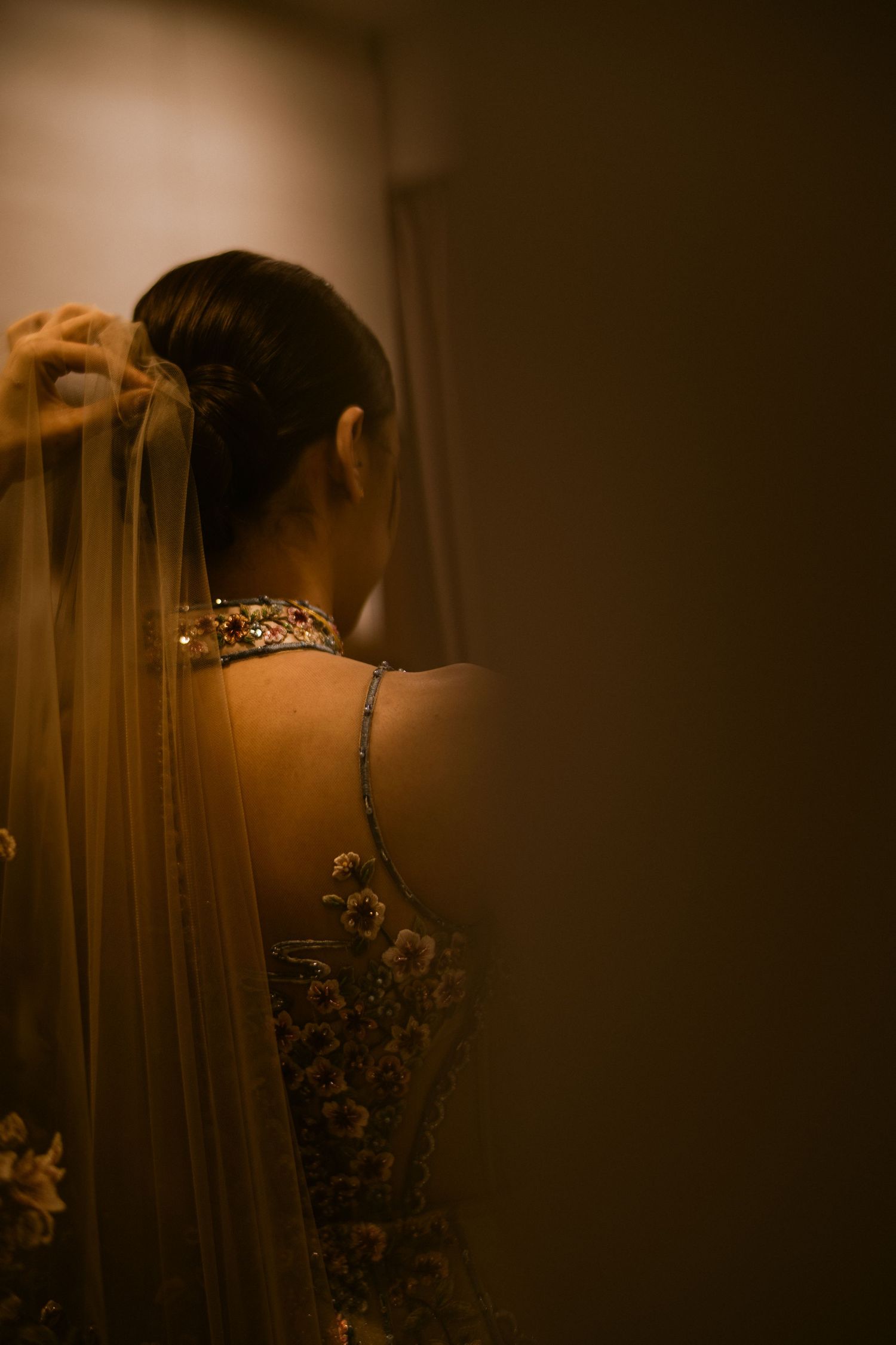Bride in ornate embellished gown with veil being placed, viewed from behind in warm golden light.