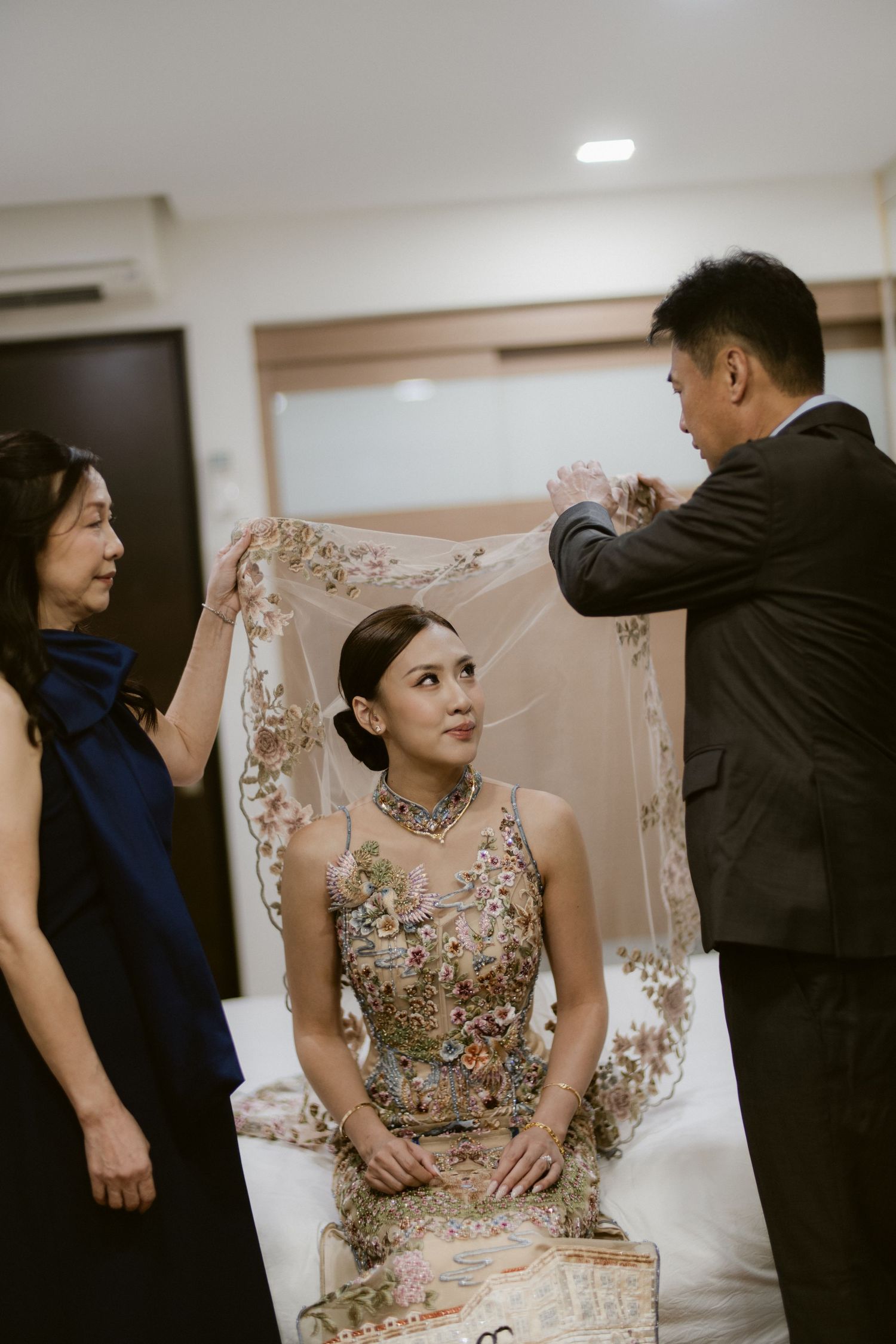 Bride seated in ornate embroidered gown as parents place veil during pre-wedding getting ready moment.