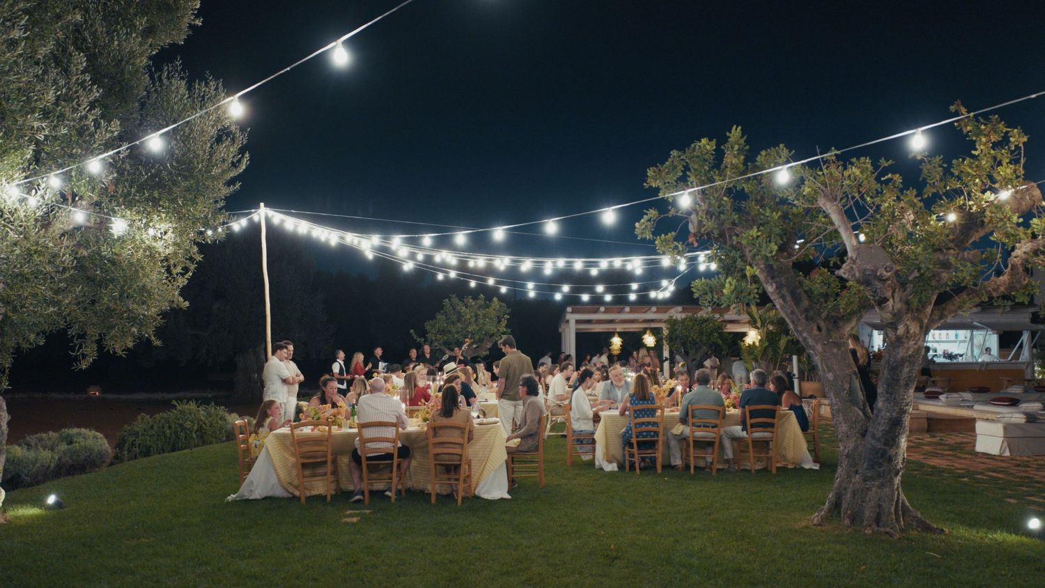 Elegant outdoor evening wedding reception with string lights, guests dining at long tables beneath olive trees in a garden.