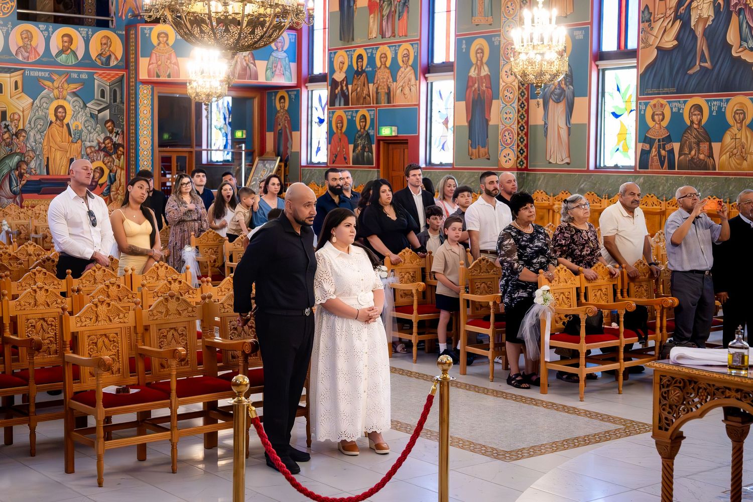 Guests gathered inside an ornate Orthodox church with golden iconostasis during a religious ceremony.
