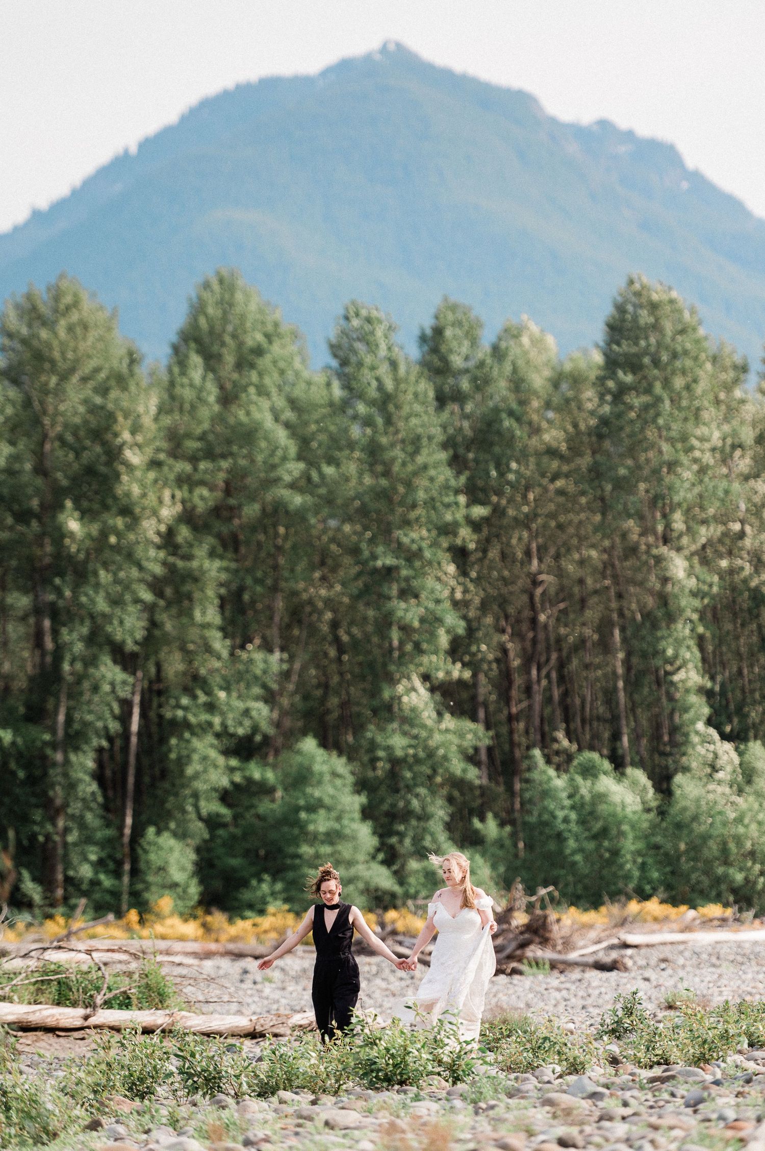 Two brides in front of a scenic mountain landscape with pine trees and a rocky riverbank in the foreground during their Packwood elopement