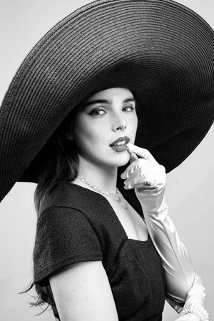 Girl wearing a big black hat,  blue pencil dress and pink elbow-length gloves, looking straight with finger in mouth, black and white photo