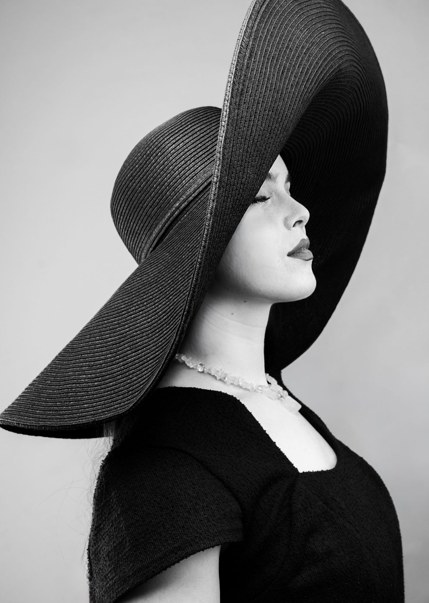 Black and white side profile photo fo a girl with pearls, a big black hat, looking right with eyes closed