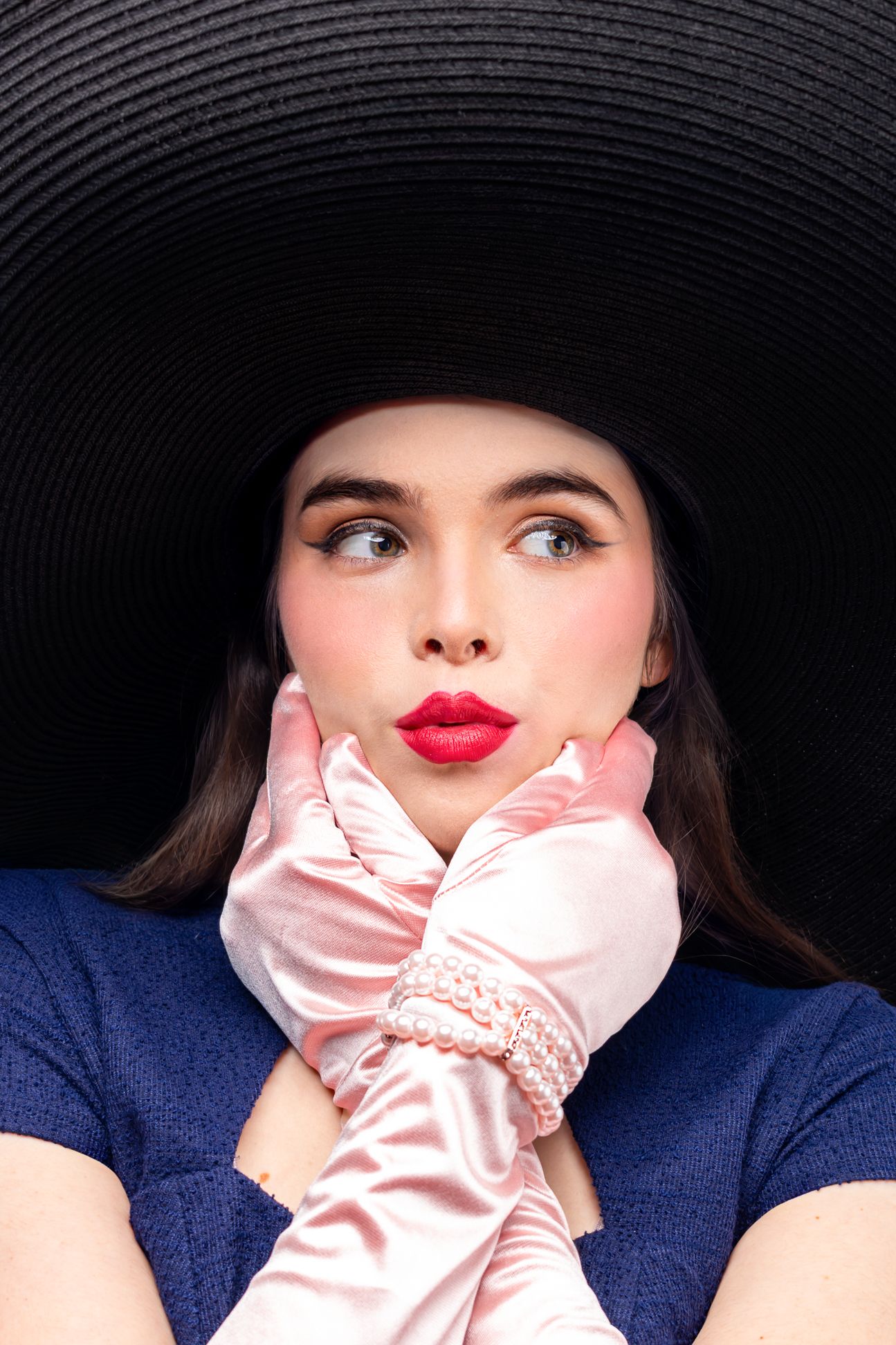 Girl wearing a big black hat, blue pencil dress and pink elbow-length gloves, hands touching the face with lips pursed, looking right