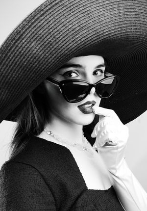 Black and white photo of a girl wearing a big black hat, big black glasses and gloves, looking at camera with glasses slightly down