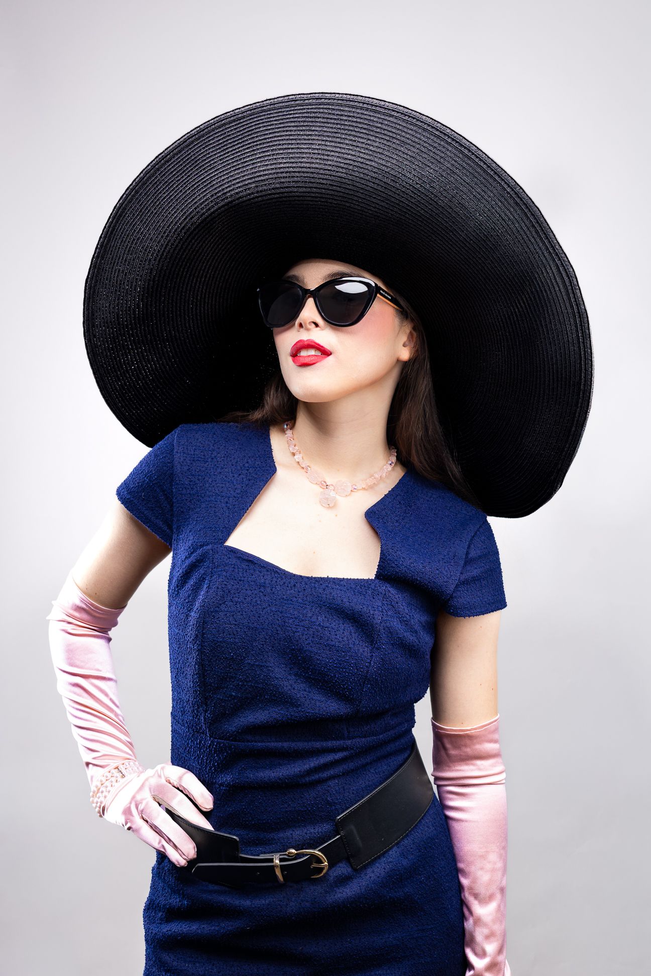 Girl wearing a big black hat, black sunglasses, blue pencil dress and pink elbow-length gloves, hand on hip, looking up to the right