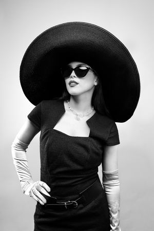 Girl wearing a big black hat, black sunglasses, blue pencil dress and pink elbow-length gloves, hand on hip, looking up to the right, b/w