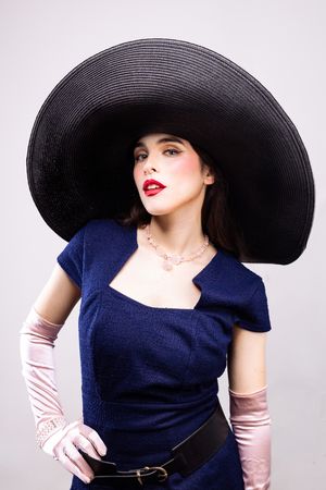Girl wearing a big black hat,  blue pencil dress and pink elbow-length gloves, hand on hip, looking straight