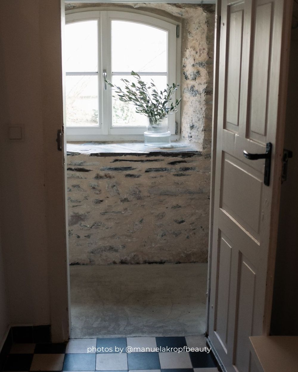 Interior hallway with white doors and distressed walls showing exposed brick and plaster near a bright window.