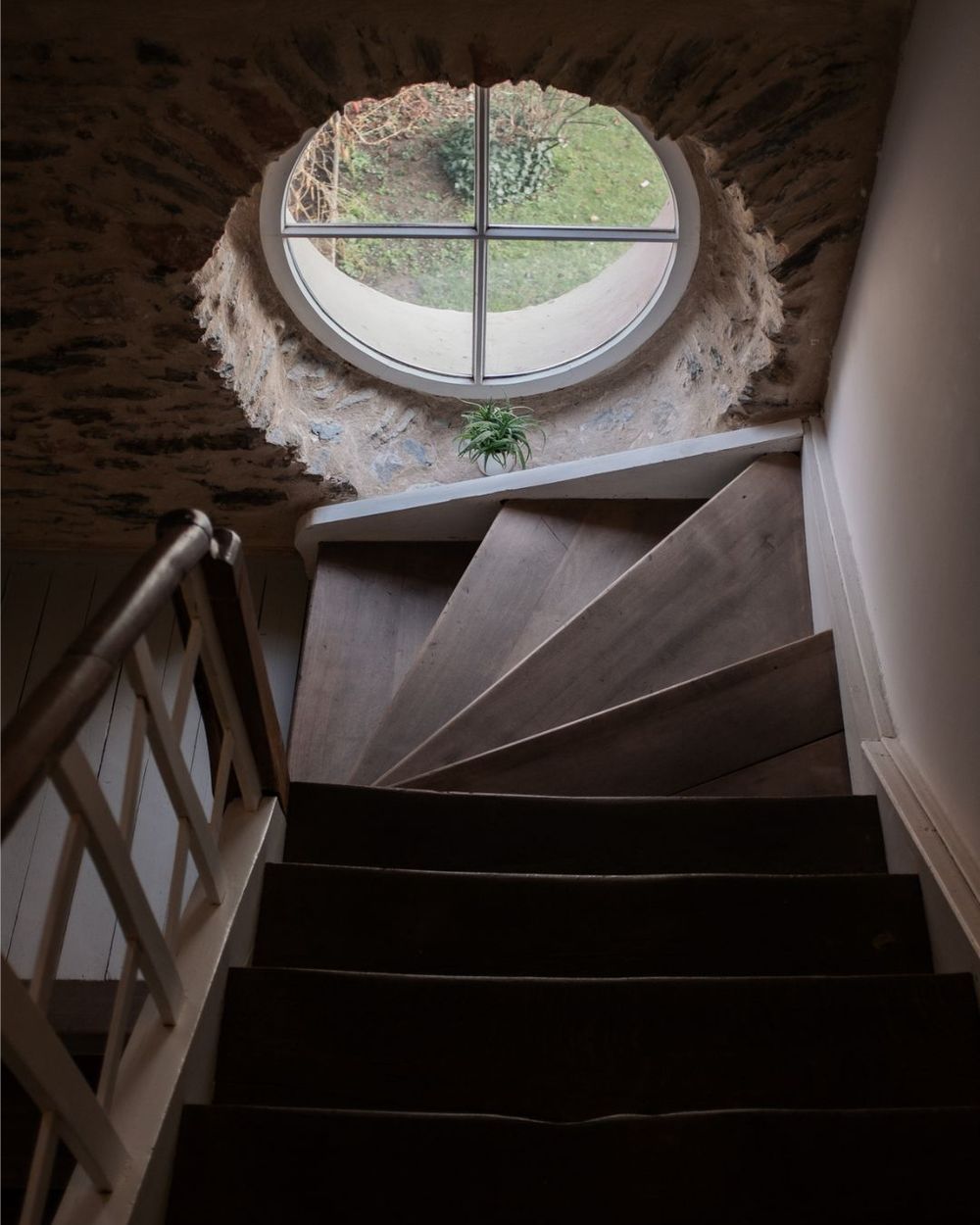 View up a dark spiral staircase with a circular window at the top letting in natural light through weathered plaster walls.
