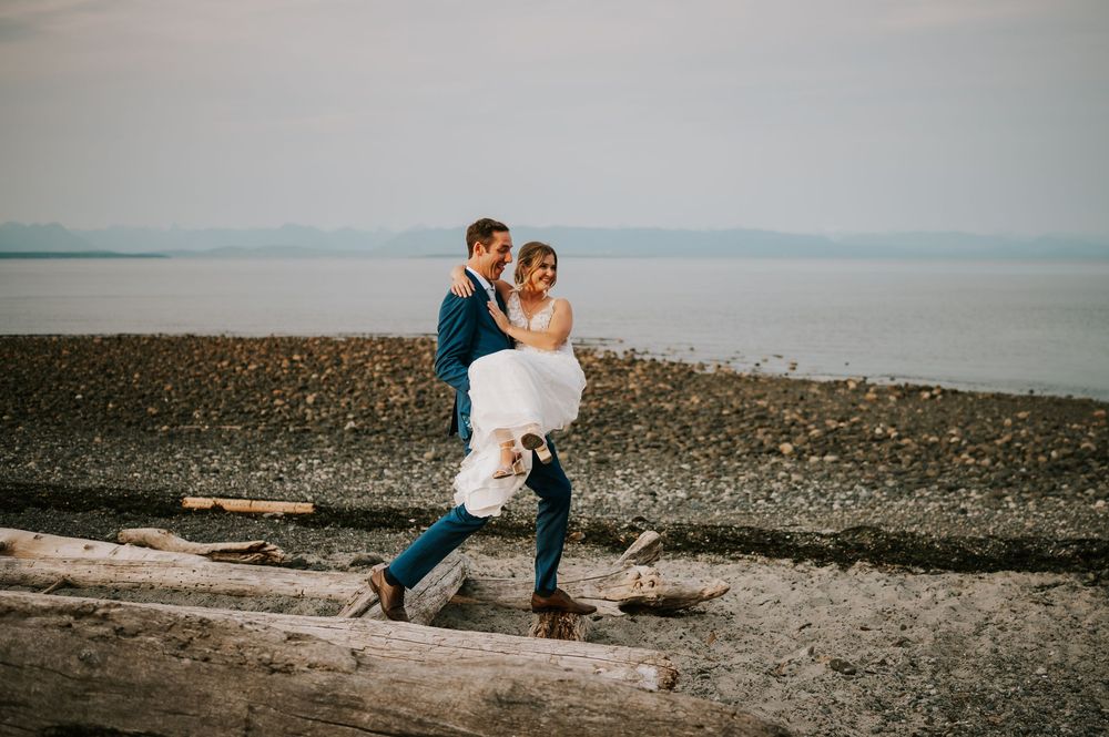 Wedding photography of couple in Merville on Vancouver Island Beach, groom carrying bride with ocean and mountains in background