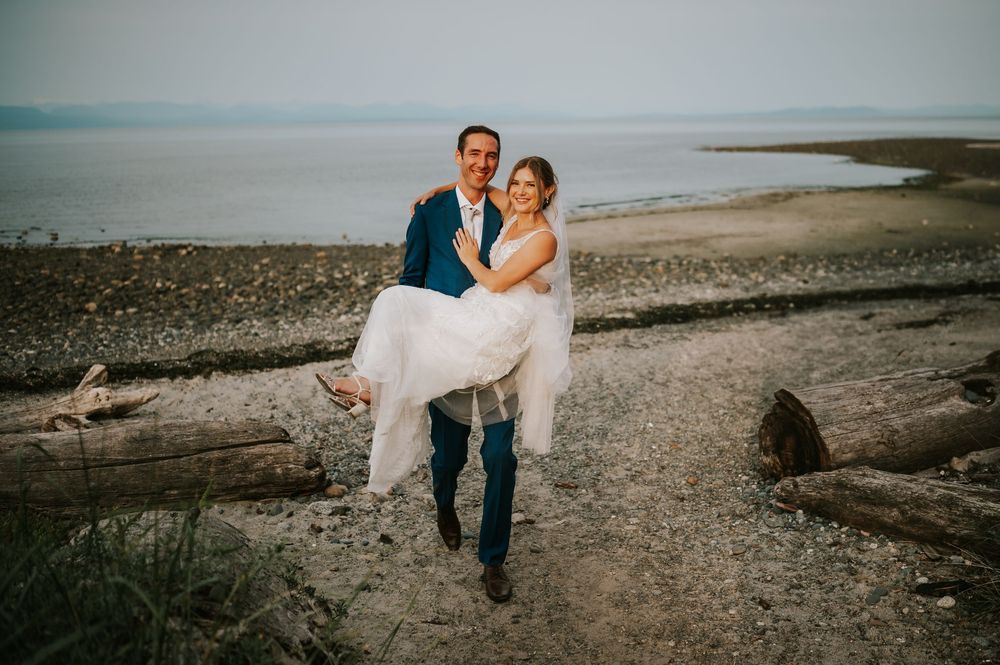 Wedding photography of couple in Merville on Vancouver Island Beach, groom carrying bride with ocean and mountains in background