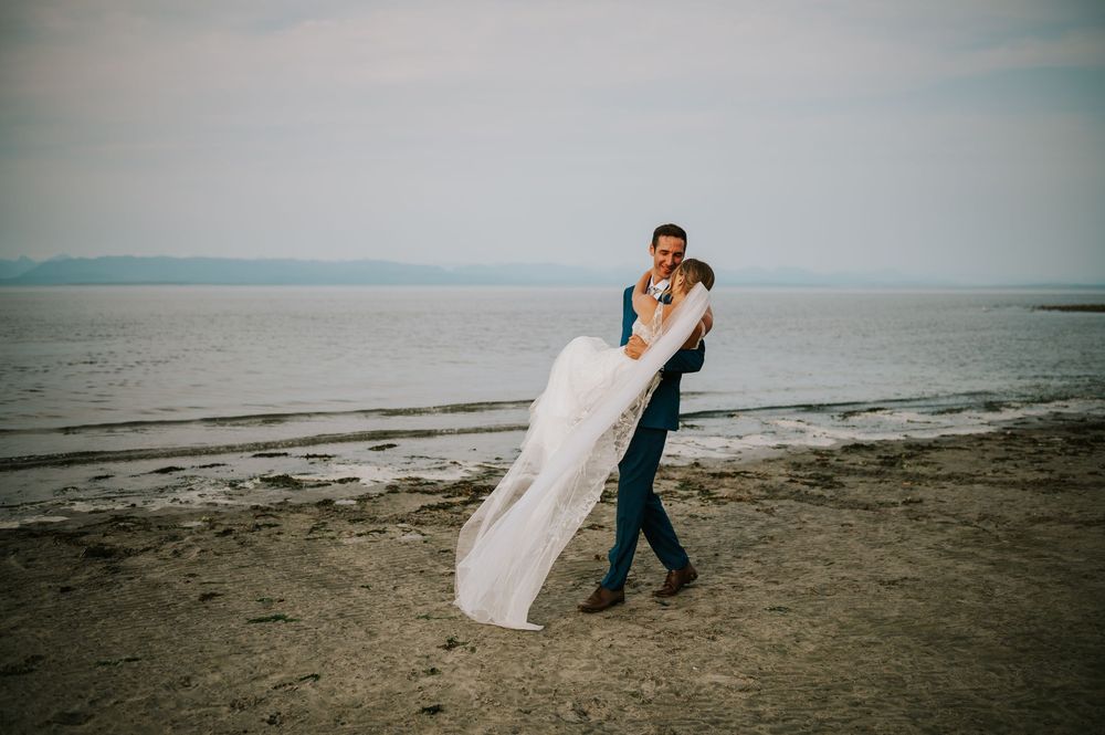 Wedding photography of couple on Williams Beach, groom carrying bride during golden hour with ocean, and mountains in background