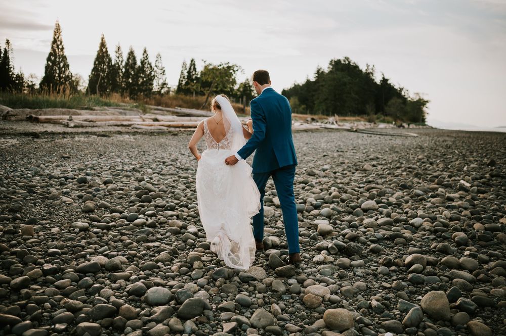Wedding photography of couple on Williams Beach, groom carrying brides wedding dress on rocky beach during golden hour