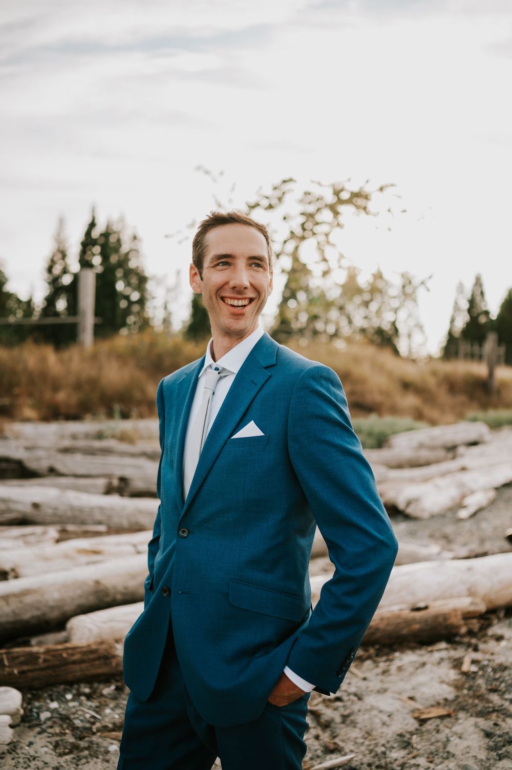 Groom smiling at ocean with driftwood and golden grass in the background, wedding photography of couple on Williams Beach