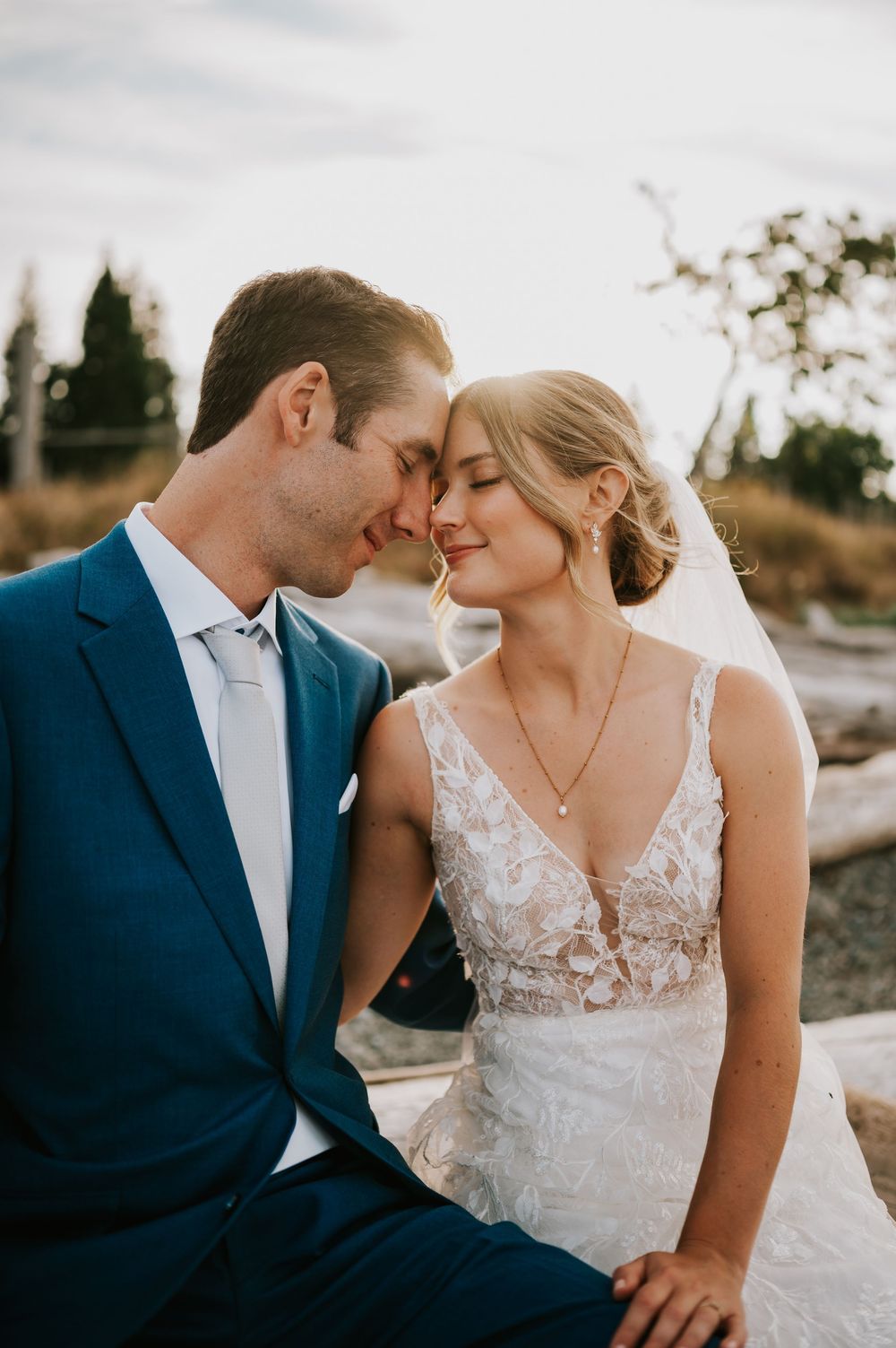 Close of portrait of bride and groom sitting on driftwood with foreheads touching, wedding photography of couple on Williams Beach