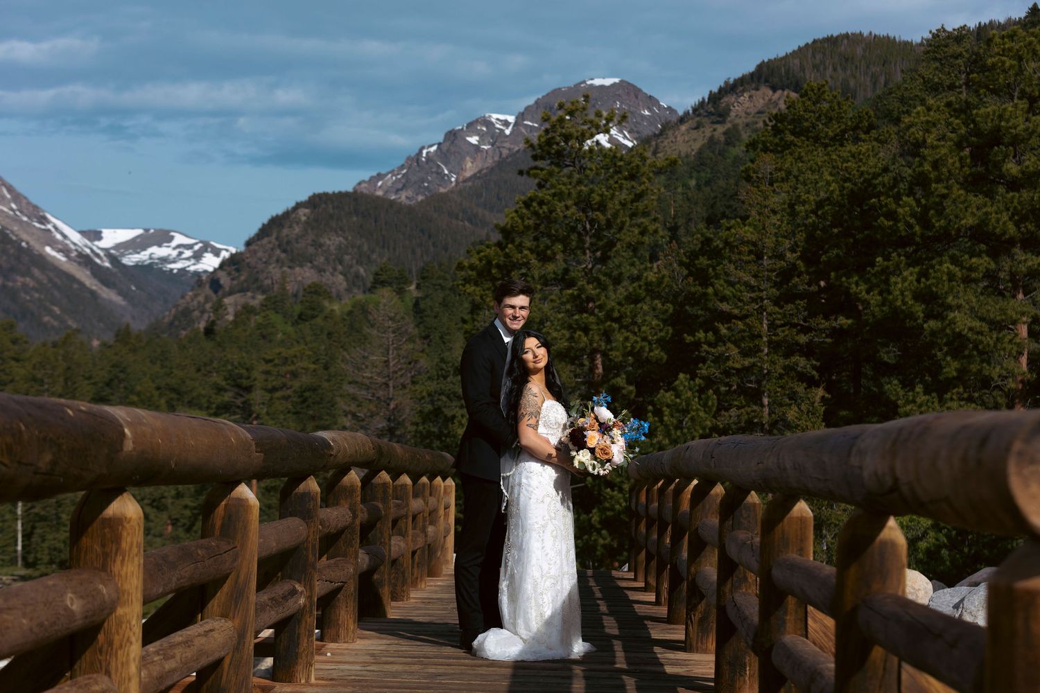 A bride and groom pose on a wooden bridge surrounded by snow-capped mountains and evergreen forest in Rocky Mountain National Park.