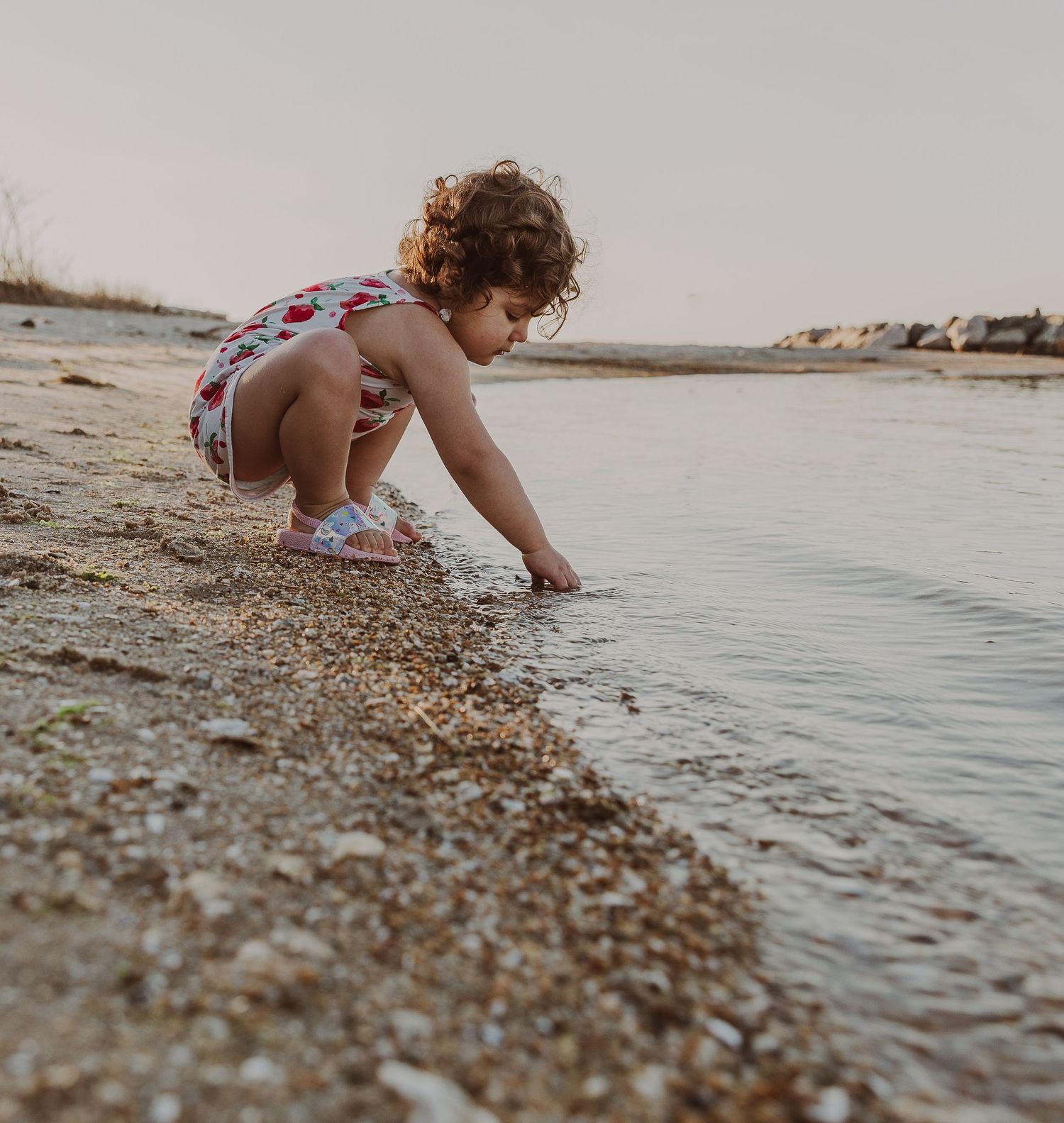 A young curly-haired girl crouches at the water's edge on a sandy beach at sunset.