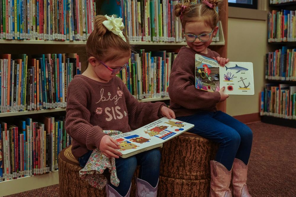Two young girls reading colorful books together in a library, sitting on tree stump stools among shelves of children's books.